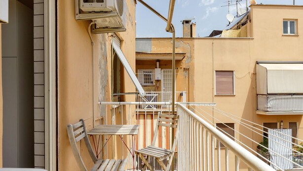 A balcony with a wooden chair and a table, a white railing, and a building with peeling paint and satellite dishes. - Apartment 'Amy Home' with Balcony, Wi-Fi and Air Conditioning (Roma)