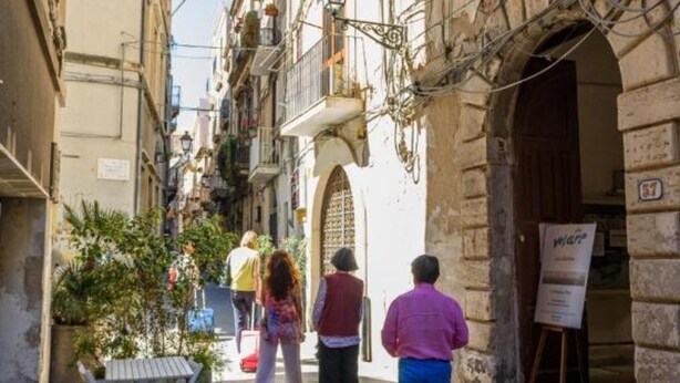 A narrow street with outdoor seating, stone buildings, and a signboard.