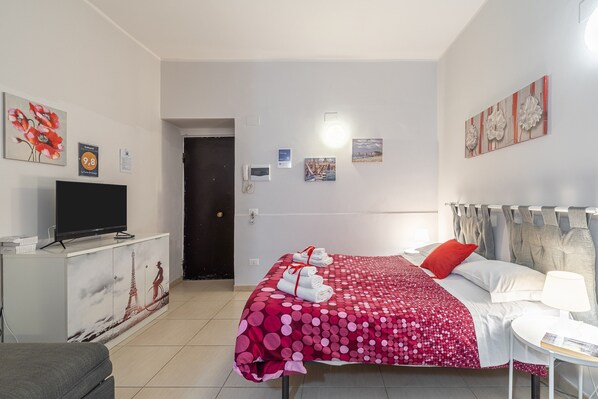 A hotel room with a bed, a TV on a cabinet, a desk with a lamp, and artwork on the walls.