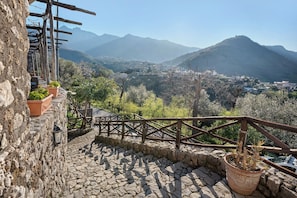Una escalera de piedra conduce a una terraza con barandilla de madera, con vistas a un valle con edificios y montañas.