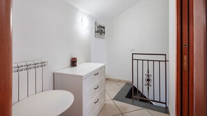 Ein Zimmer mit einem weißen Heizkörper, einer weißen Kommode mit Schubladen, einem runden Tisch, einem schwarzen Metallgitter und einer weißen Wand mit einer Leuchte.