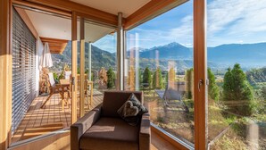 Ein modernes Zimmer mit einer großen Glasschiebetür, einer Holzterrasse, einer Couch mit Kissen und Blick auf Berge und ein Tal.