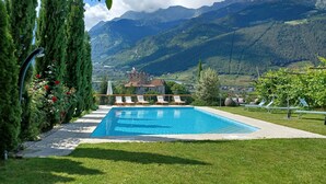 Ein Schwimmbecken mit Liegestühlen, umgeben von Grünflächen und Bergen im Hintergrund.