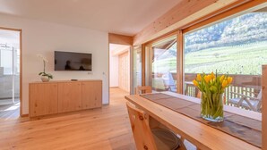 Un intérieur moderne avec une table à manger en bois, un téléviseur fixé au mur et une vue sur un vignoble grâce à de grandes fenêtres.