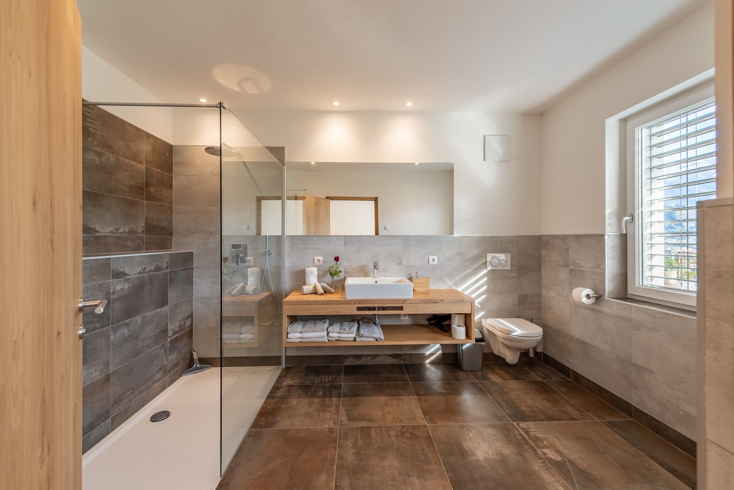 A modern bathroom with a glass shower enclosure, a wooden vanity, a toilet, and a large window.