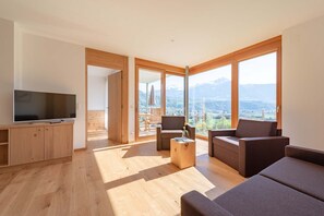 Ein modernes Wohnzimmer mit Flachbildfernseher, Holzmöbeln und großen Fenstern mit Blick auf die Berge.