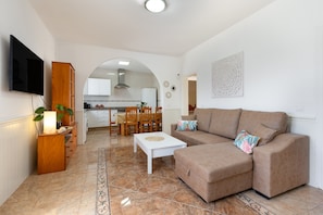 A living room with a sofa, a TV mounted on the wall, a dining area with chairs and a table, and a kitchen in the background.