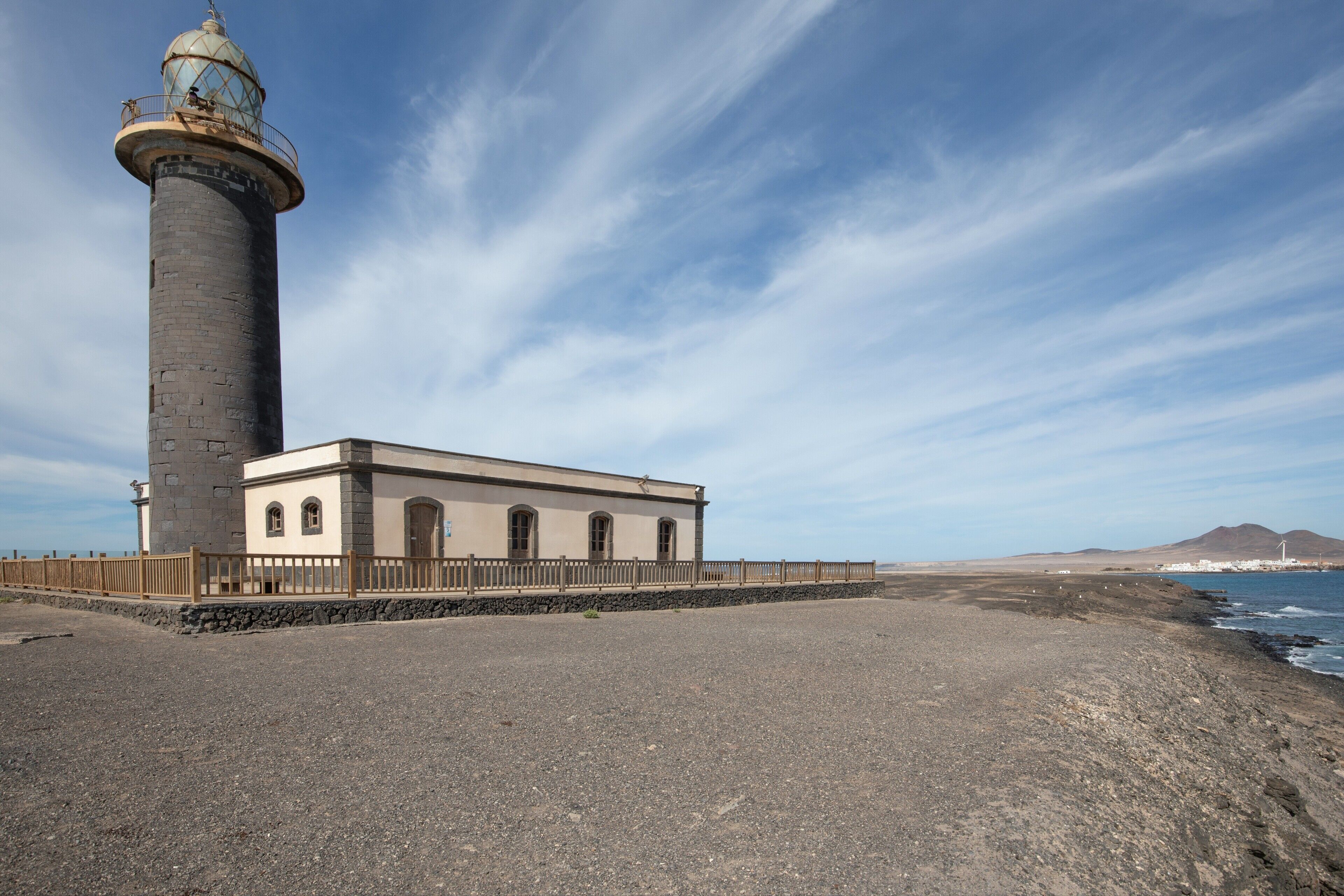 Un faro con torre de piedra y un edificio con ventanas de arco, situado en una playa rocosa bajo un cielo despejado.