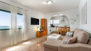 A living room with a sofa, a coffee table, and a view of the sea through a window with sheer curtains. - Vacation Home 'Buganvilla' with Sea View and Wi-Fi (Pájara)