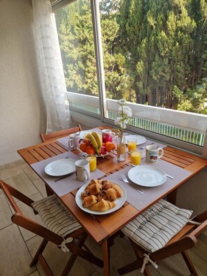 A breakfast table with plates, croissants, fruit, and juice, set on a balcony with a view of trees. - Studio 'Sanary Sur Mer, Soleil Et Douceur I' with Private Terrace, Wi-Fi and Air Conditioning (Sanary-sur-Mer)