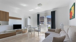 A modern living room with a flat-screen TV, a sofa, a dining table with chairs, and a large window with curtains. - Escapada Al Sur Vacation Apartment with Wi-Fi (Morro del Jable)