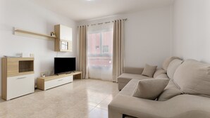 Ein modernes Wohnzimmer mit einem Flachbildfernseher, einem beigefarbenen Sofa, einem Holzschrank und einem Fenster mit Vorhängen.