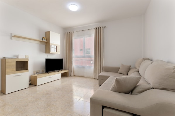 Una sala de estar moderna con un televisor de pantalla plana, un sofá color beige, un mueble de madera y una ventana con cortinas.