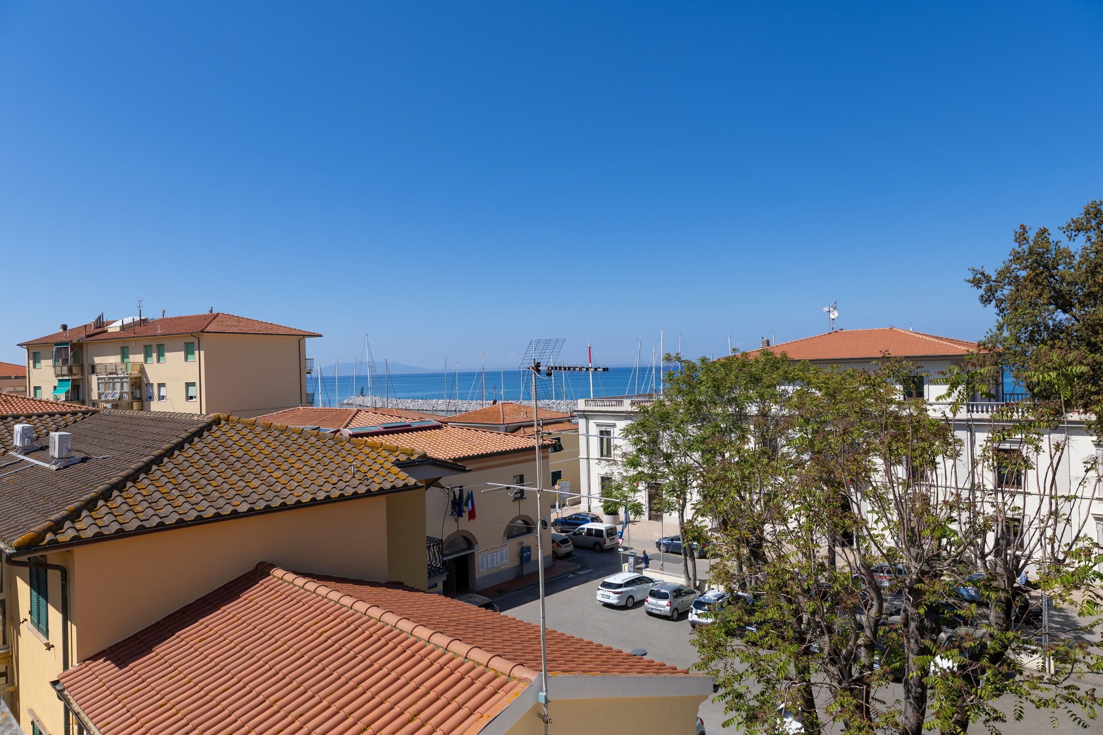 A coastal town with terracotta-roofed buildings, a clear blue sky, and a view of the sea.