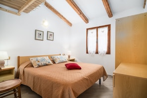 Un dormitorio con una cama de madera, una mesita de noche, una silla, una ventana con cortinas y vigas de madera en el techo.