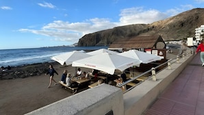 A coastal area with outdoor seating under umbrellas, a rocky beach, and a hillside in the background. - Vacation 'Dream Home Beny' with Sea View, Wi-Fi and Air Conditioning (Palm-mar)