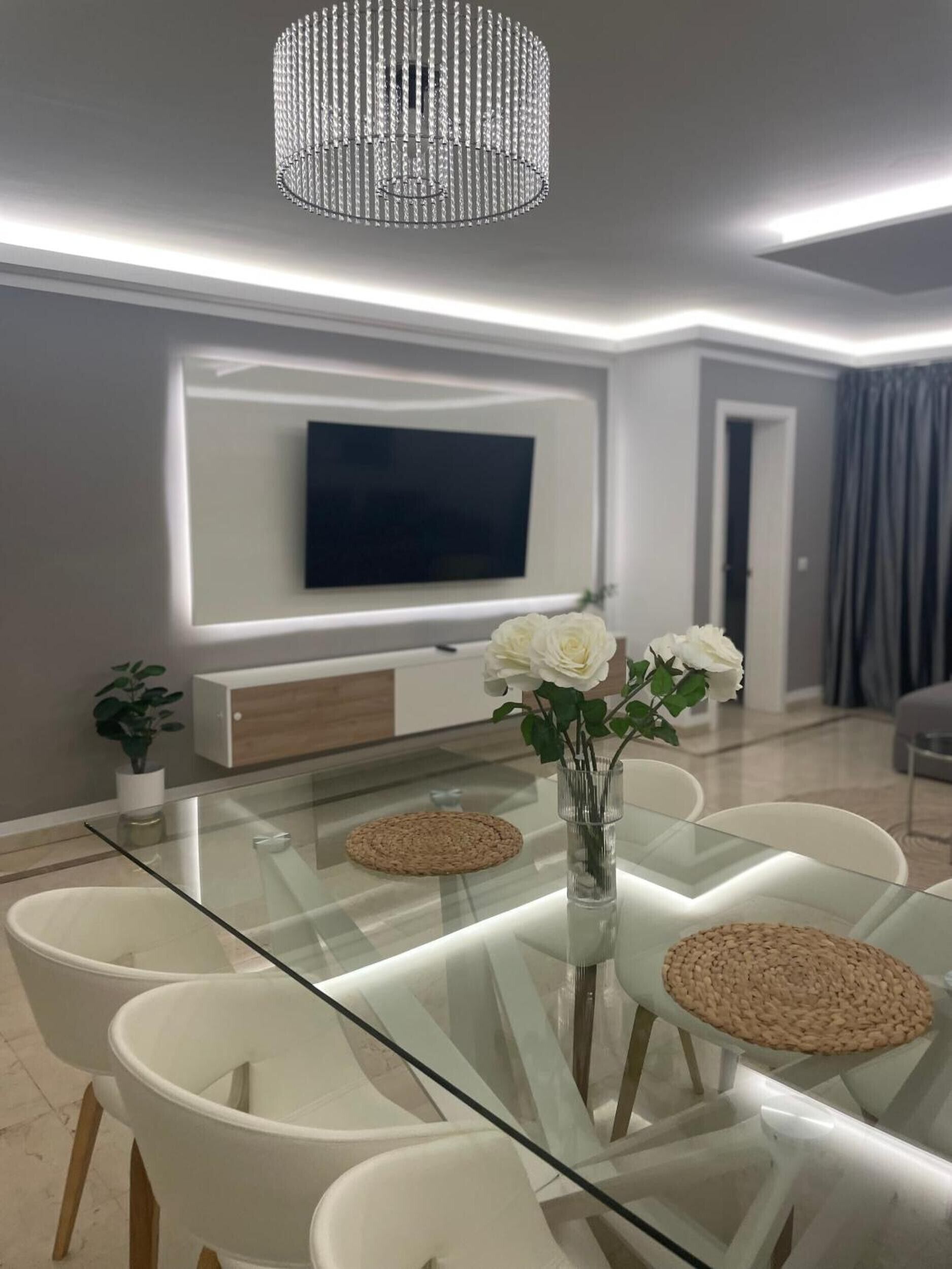 Un comedor moderno con una mesa de vidrio, sillas blancas, un jarrón con flores blancas, un televisor montado y una lámpara decorativa.