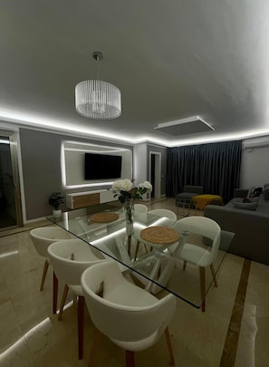 Un salon moderne avec une table à manger en verre, des chaises blanches, un lustre, un téléviseur à écran plat et un canapé.