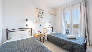 A hotel room with two beds, a small table with a flower, a framed botanical print on the wall, and a balcony with a view.
