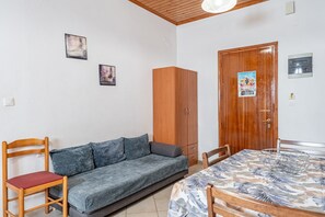 Um quarto com sofá, uma cadeira de madeira, uma mesa de jantar com toalha de mesa estampada com folhas, um guarda-roupa de madeira e duas molduras na parede.
