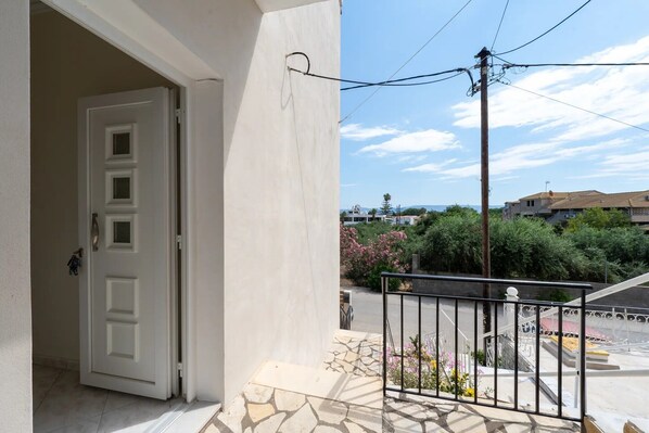 Une porte ouverte donnant sur un balcon doté d’un sol en pierre, d’une rambarde en métal et offrant une vue sur un quartier résidentiel avec des arbres et des immeubles.
