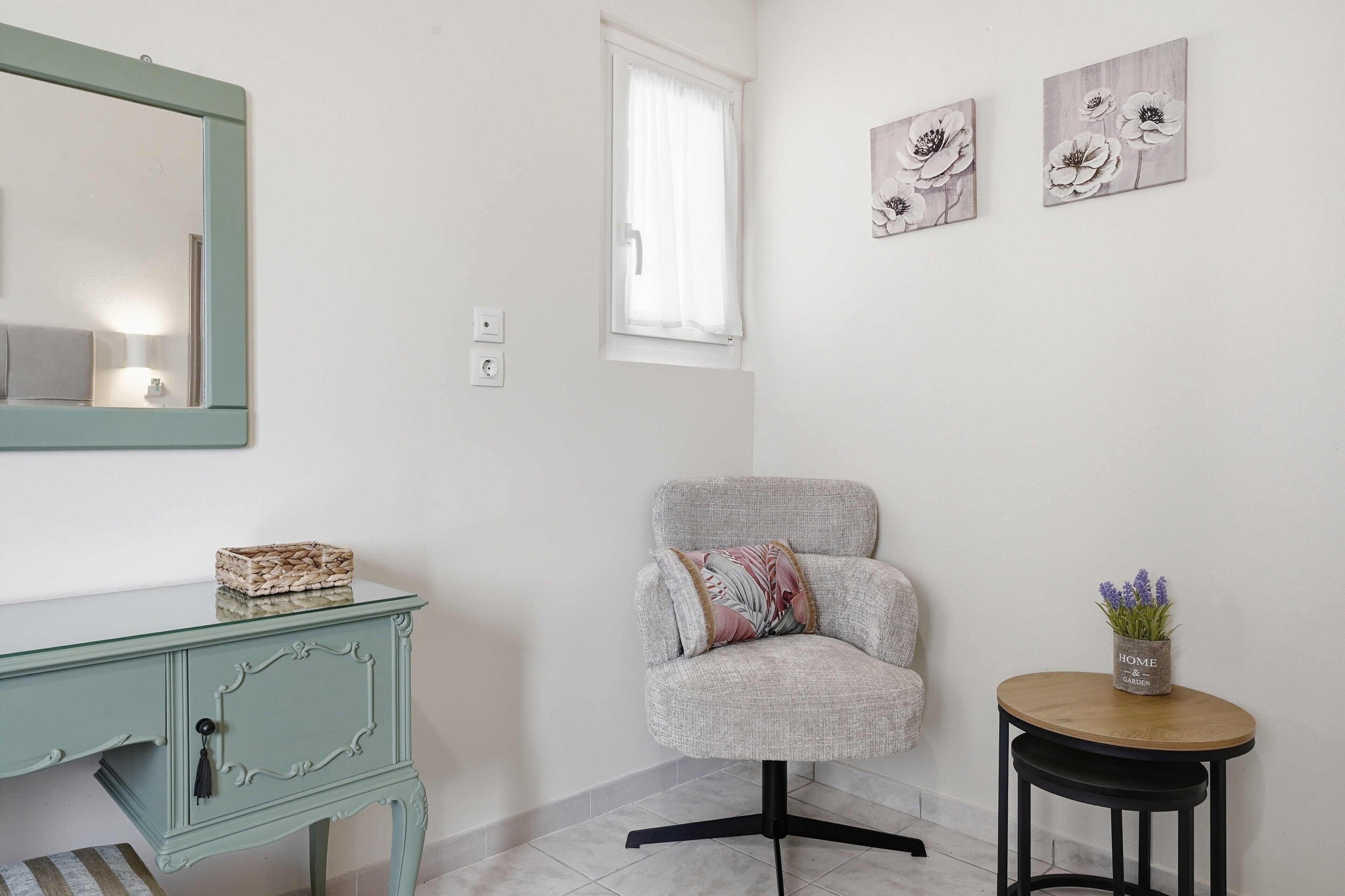 Une pièce avec une armoire à portes miroir, un fauteuil gris avec un coussin, une petite table ronde en bois sur laquelle est posée une plante en pot, et deux tableaux représentant des fleurs, accrochés au mur.