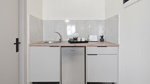 A compact kitchen with white cabinetry, a built-in dishwasher, and a countertop with a kettle, cups, and a small plant.