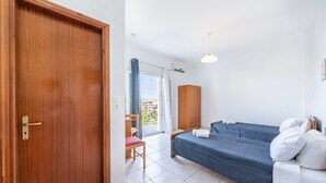 A bedroom with a wooden door, a bed with a blue mattress, a wooden wardrobe, a chair, and a view through a window.