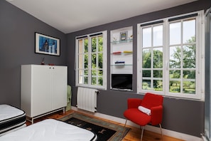 A room with a bed, a red chair, a white wardrobe, shelves, and large windows with a view of greenery.