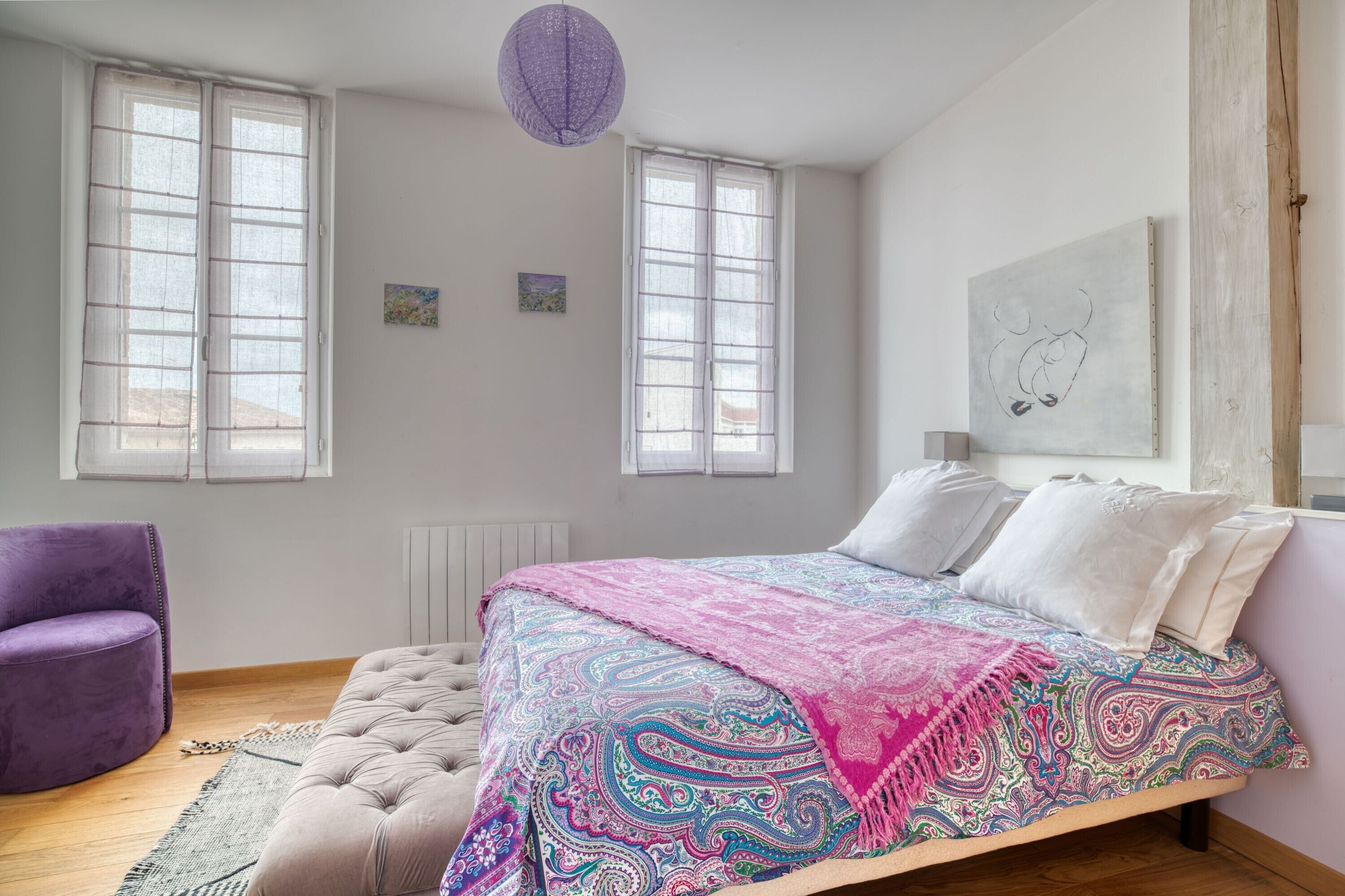 Une chambre à coucher avec un lit, deux fenêtres, un fauteuil violet et une œuvre d’art murale.