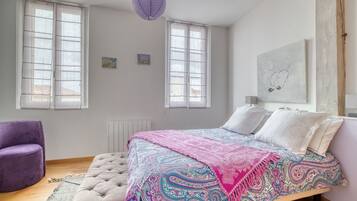 Une chambre à coucher avec un lit, deux fenêtres, un fauteuil violet et une œuvre d’art murale.