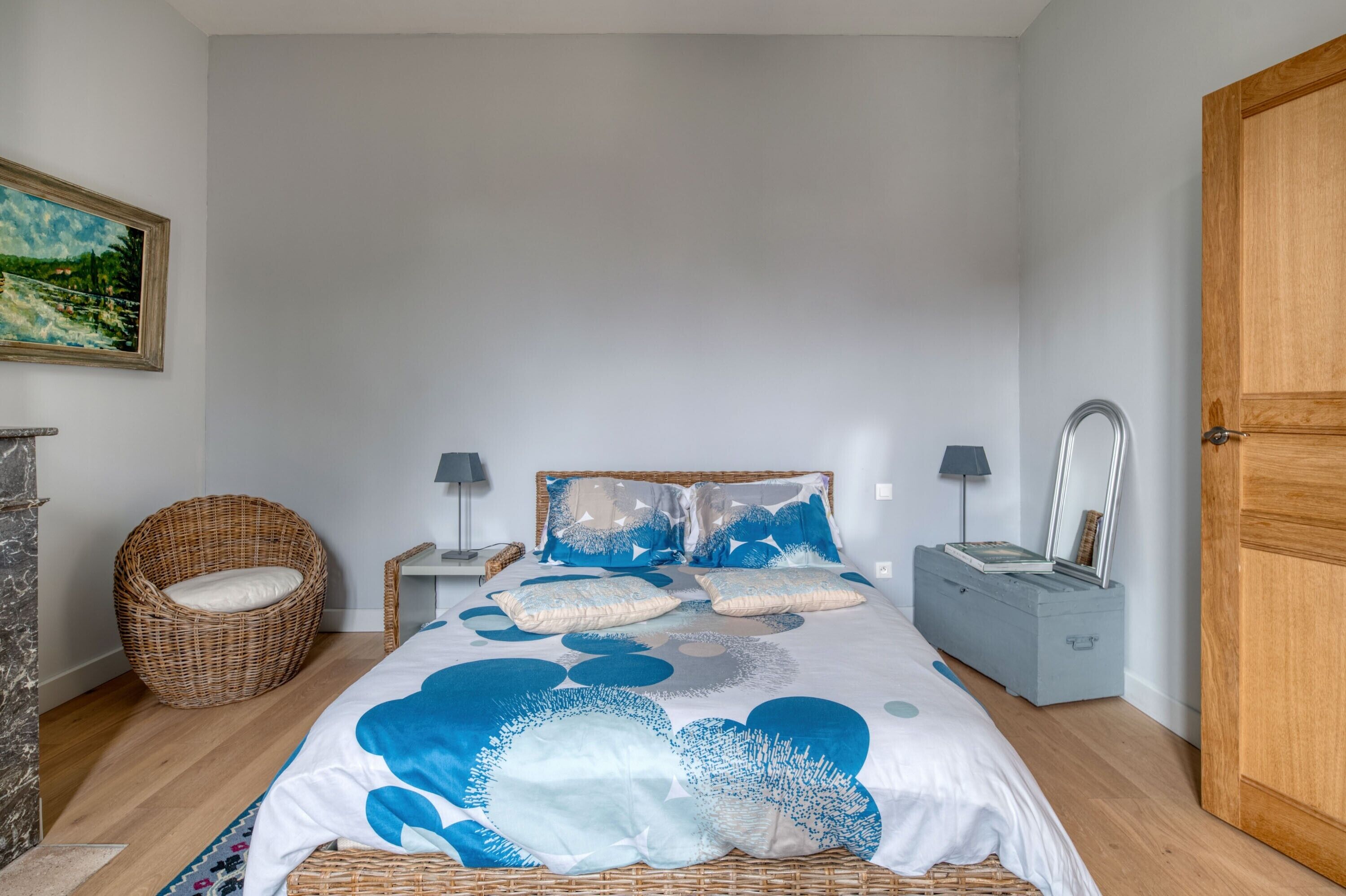 Ein Schlafzimmer mit einem Bett, einem Korbstuhl, einem Nachttisch, einer Lampe, einer Kommode und einer Tür.