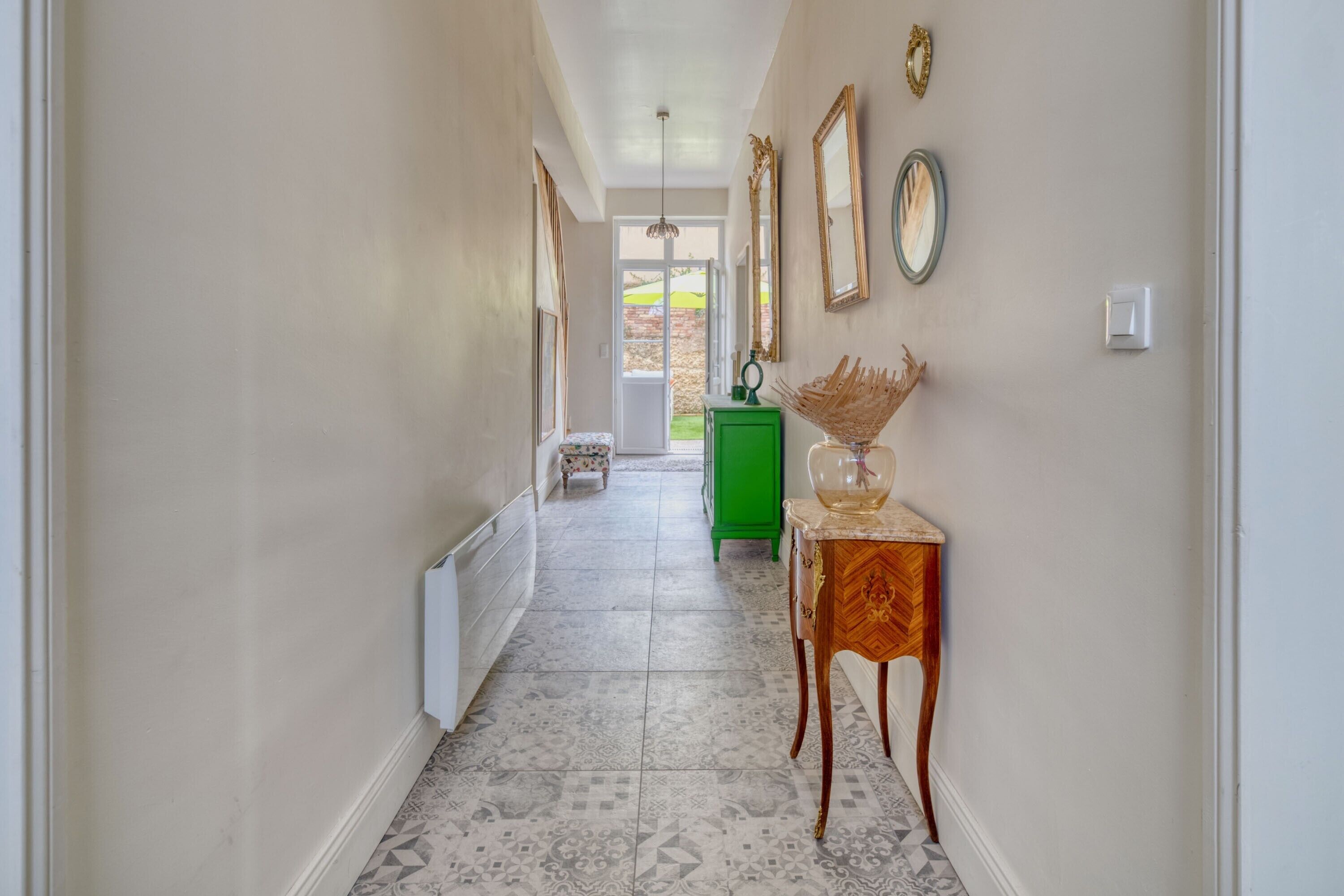 Un couloir avec des carreaux de sol à motifs, un meuble vert et une table décorative sur laquelle est posé un objet en verre.