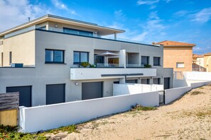 A modern two-story building with a flat roof, large windows, and a balcony with outdoor furniture.