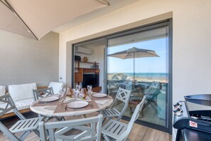 A balcony with a table set for dining, a grill, and a view of the beach.