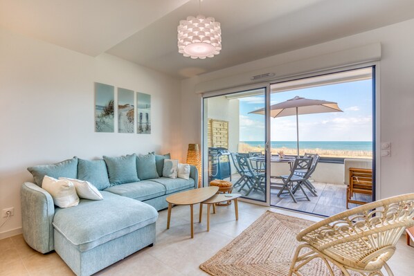 A living room with a sofa, coffee table, and wicker chair, a dining area with chairs and a table, and a large sliding glass door leading to a balcony with an umbrella.