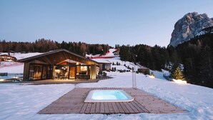 A wooden deck with a hot tub in a snowy landscape, a building with large windows, and a mountain in the background. - Chalet 'Ski' on the slopes with mountain view, private terrace, and Wi-Fi (Selva di Val Gardena)