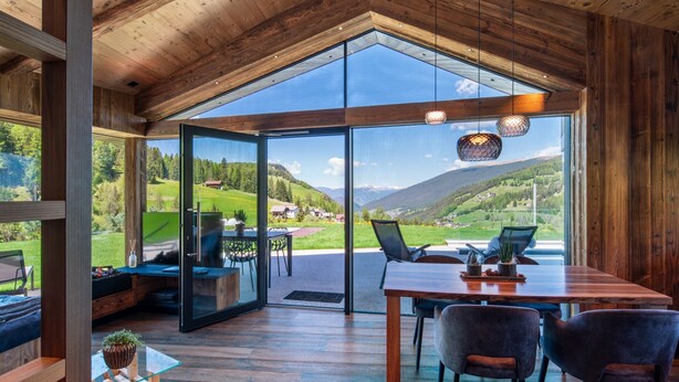 A modern interior with a wooden ceiling, a glass door leading to a scenic outdoor view, a dining area with chairs and a table, and a small seating area with a sofa and a plant. - Chalet 'Ski' on the slopes with mountain view, private terrace, and Wi-Fi (Selva di Val Gardena)