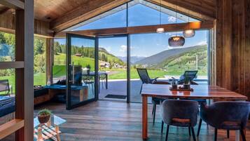 Interior moderno com teto de madeira, porta de vidro que dá para uma vista deslumbrante do exterior, área de jantar com cadeiras e mesa, e um pequeno espaço de estar com sofá e planta.