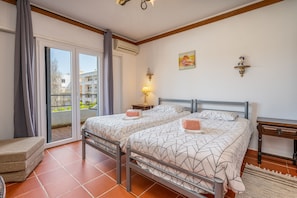A bedroom with two beds, a sliding glass door to the balcony, a small bedside table, and a wall-mounted lamp.