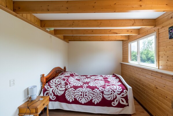 Un dormitorio con techo de madera, una cama con colcha estampada, una mesita de noche con lámpara y una ventana con vistas al verdor.