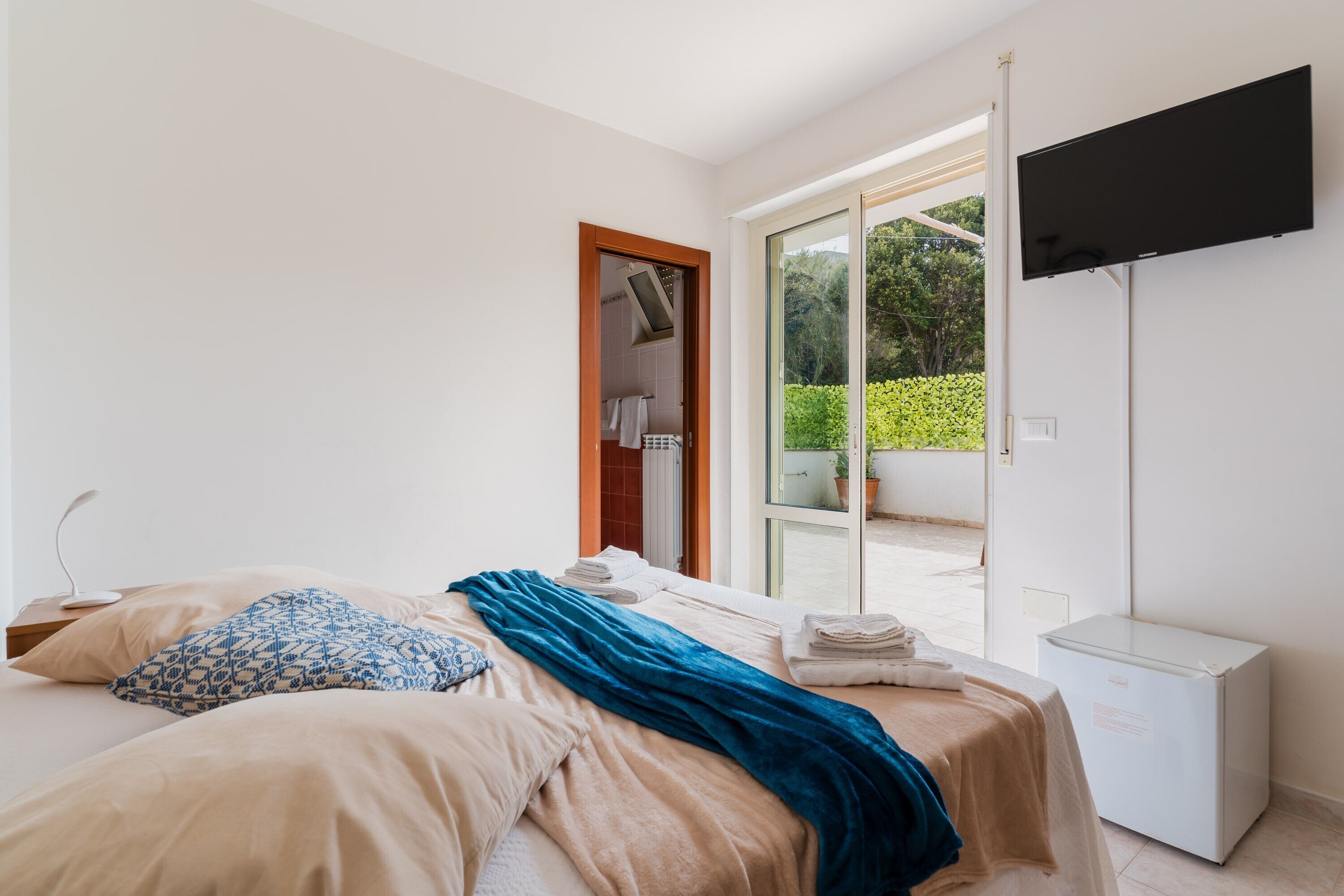A bedroom with a bed, a TV, a mini-fridge, and a sliding glass door to a balcony.