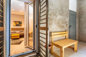 Una habitación con un banco de madera, un ventanal corredero que da a una cama con un reposapiés amarillo y una pared de concreto.