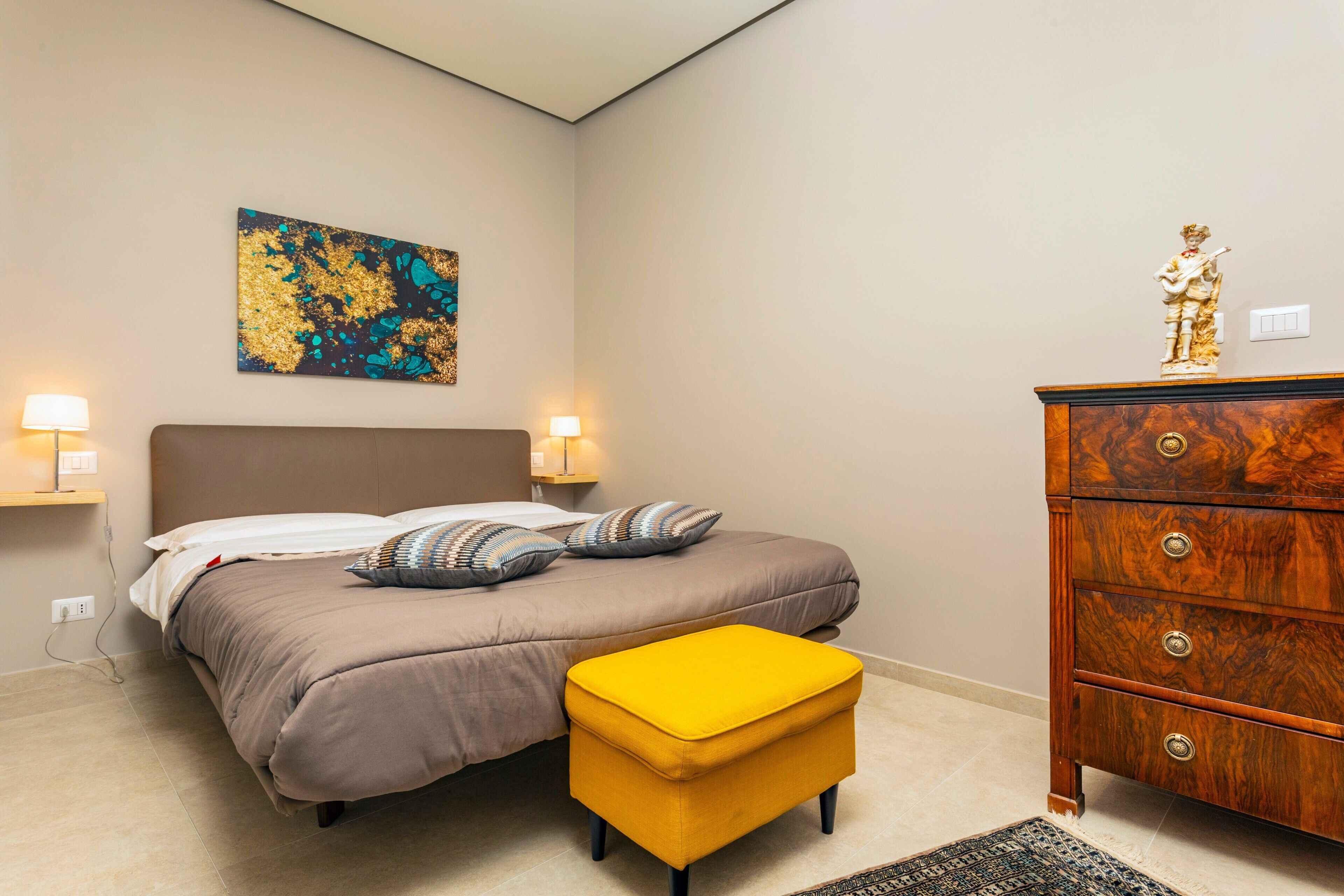 Ein Schlafzimmer mit einem Bett, Nachttischlampen, einer Holzkommode, einem farbenfrohen abstrakten Gemälde und einem kleinen, gelben Hocker.