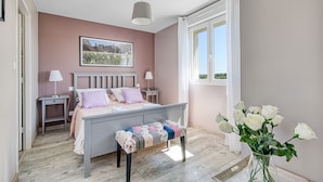 Une chambre à coucher avec un lit, des tables de chevet, un banc, une fenêtre et un vase rempli de fleurs.