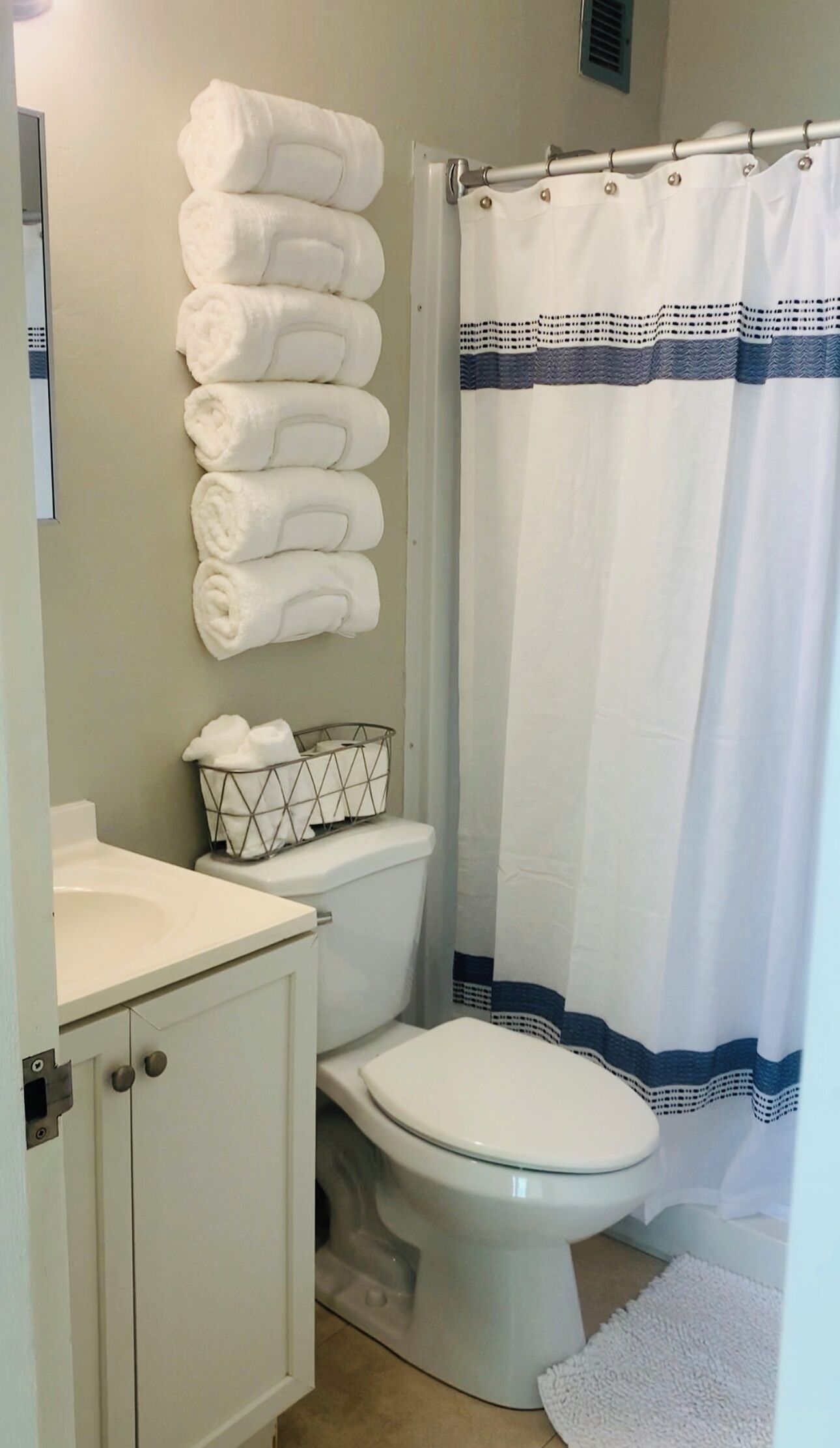 Une salle de bain comprenant des toilettes, un lavabo, un porte-serviettes avec des serviettes pliées, un rideau de douche et un petit panier posé sur le lavabo.