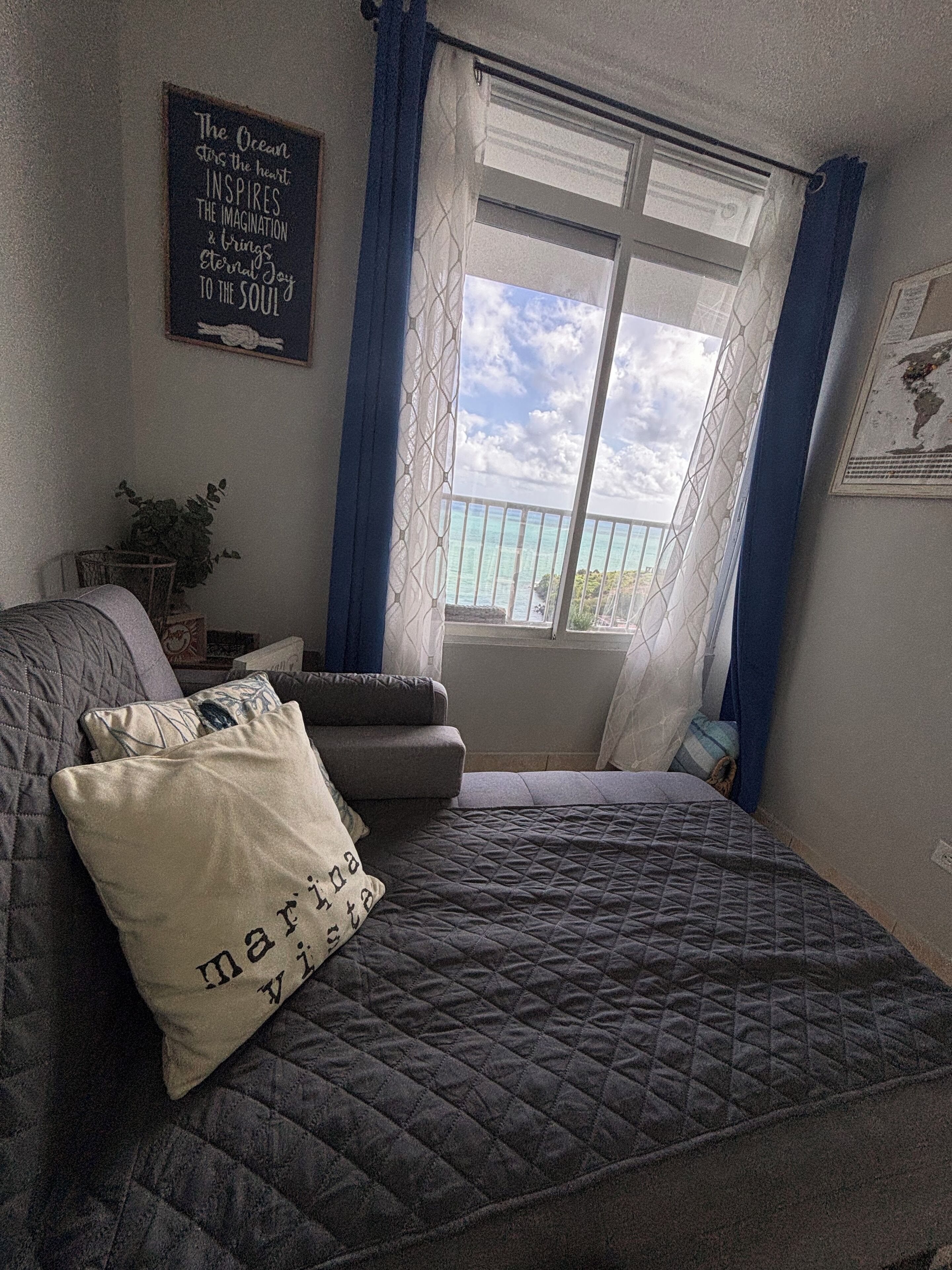Un dormitorio con cama, ventana con cortinas, pared decorada con una cita enmarcada y una mesita con planta.