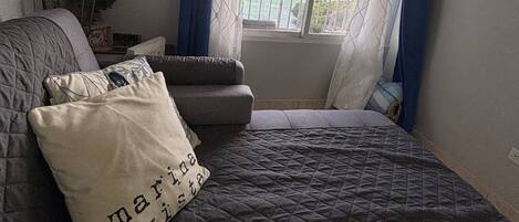 Un dormitorio con cama, ventana con cortinas, pared decorada con una cita enmarcada y una mesita con planta.