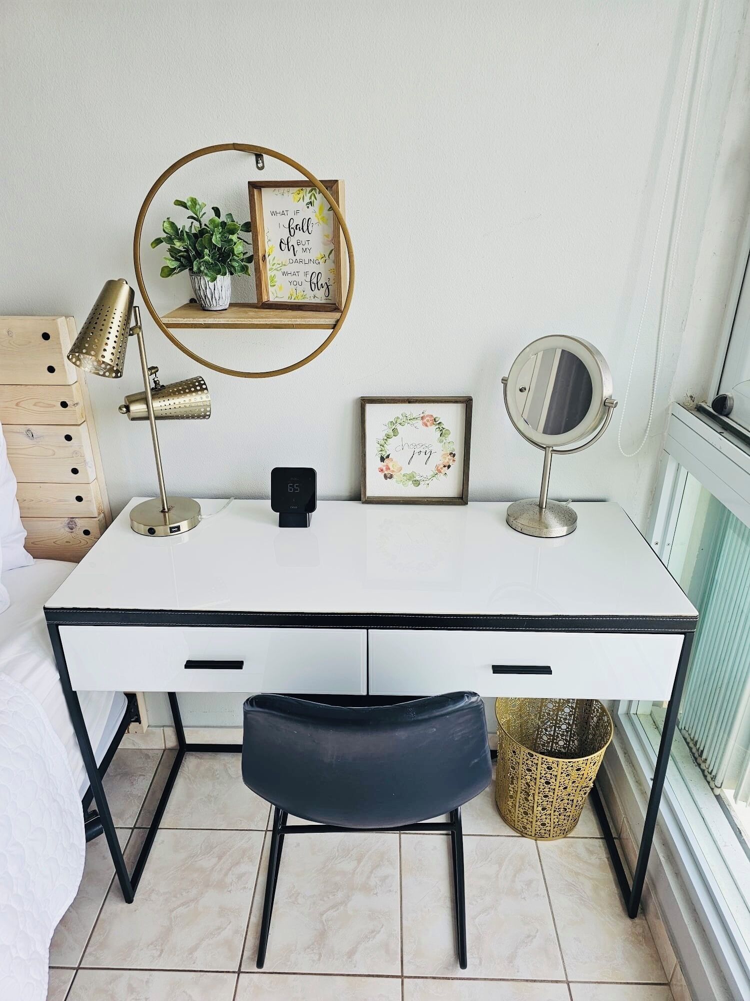 Un bureau blanc avec deux tiroirs, une chaise noire, un miroir rond, une lampe et un tableau encadré accroché au mur.