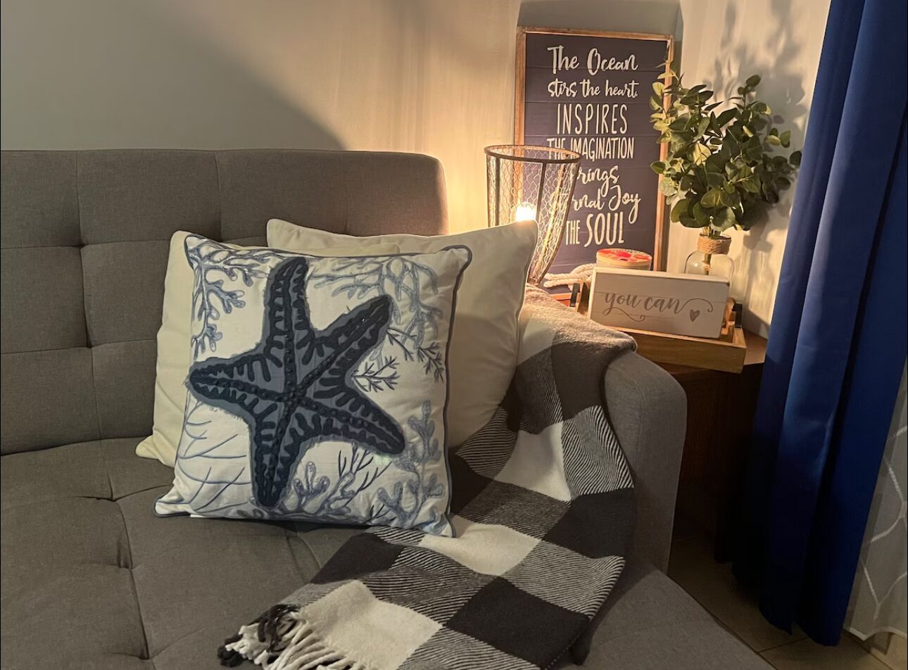 A cozy living room with a grey sofa, a starfish pillow, a checkered throw blanket, a wall-mounted sign with inspirational quotes, and a potted plant.